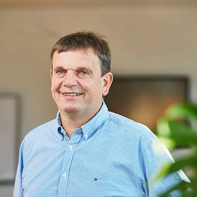 Ein Portrait von Stefan Suhr, von Endress Hauser, der lächelnd in einem sonnigen Büro hinter einer grünen Pflanze steht