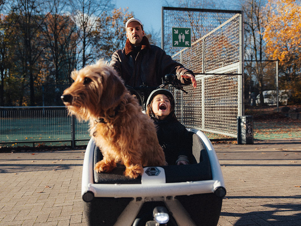 Vater mit Kind und Hund im Lastenrad mit Freudenschrei